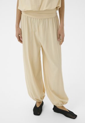 Une personne porte un pantalon beige ample avec des poignets élastiques et des chaussures à enfiler en maille noire, debout devant un fond blanc uni.