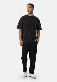 Sort bomulds T-shirt med korte ærmer, prydet med et lille blåt logo på brystet. Sammen med sorte bukser og hvide sneakers.