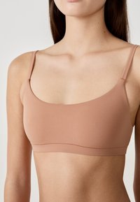 Bra marrone chiaro realizzato in un tessuto liscio e elastico. Presenta spalline sottili e un design semplice e senza cuciture, privo di decorazioni.