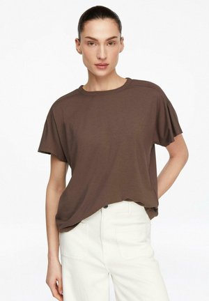 Kobieta z zaczesanymi do tyłu włosami, ubrana w luźną brązową koszulę z krótkim rękawem wpuszczoną w białe spodnie z wysokim stanem na białym tle.