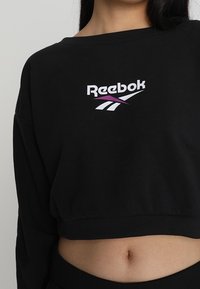 Sudadera recortada negra hecha de un tejido suave que presenta un logo de Reebok en blanco y morado en la parte delantera, con un corte relajado y un escote ancho.
