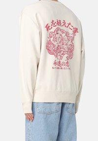 Crème-kleurige sweatshirt met lange mouwen, voorzien van een grote rode grafische opdruk op de achterkant, met schedelmotieven en Japanse tekst.