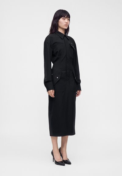 Femme aux longs cheveux foncés portant une robe midi noire à manches longues avec des détails de poches et des chaussures noires à talons hauts, debout devant un fond uni.