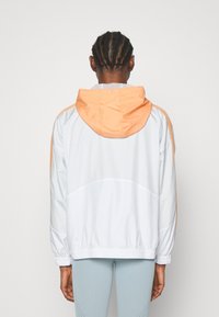 Veste coupe-vent en blanc et orange clair. Dotée d'une capuche, de poignets élastiques et d'un ourlet avec cordon de serrage. Matériau lisse et léger.