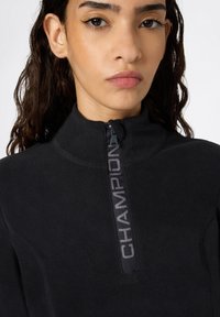 Schwarzer Fleece-Pullover mit hohem Kragen und halbseitigem Reißverschluss, versehen mit einem auffälligen grauen "CHAMPION"-Logo entlang des Reißverschlusses. Weiche Textur.