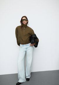 Oliwkowo-zielona krótka kurtka na zamek, jasnoniebieskie szerokie spodnie dżinsowe, czarna torebka i duże okulary przeciwsłoneczne. Gładka tkanina i luźny krój.