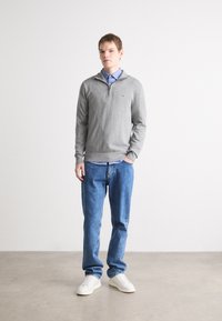 Sweatshirt zippé gris sur une chemise à col bleu, associé à un jean bleu et des baskets blanches. Design simple avec une coupe confortable.