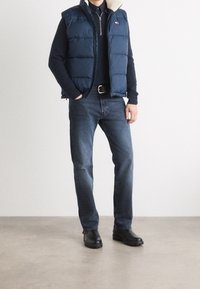 Marinblå puffer-väst med fuskpälskrage, bärs över en mörk tröja, ihop med blå jeans och svarta stövlar på en neutral bakgrund.