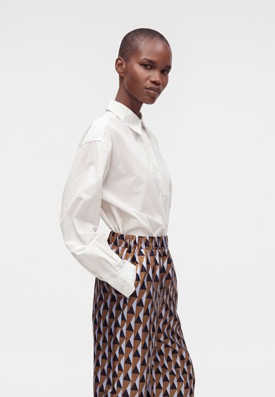 Femme aux cheveux courts porte une chemise blanche boutonnée et un pantalon taille haute avec un motif triangulaire géométrique marron, noir et blanc, mains dans les poches.