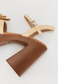 Beige hæl sandal med vævet overdel, der har en bred brun blokhæl og justerbar ankelrem med sølvhardware.