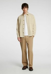 Camicia beige con bottoni e tasca sopra una camicia bianca, abbinata a pantaloni color sabbia e scarpe marroni, caratterizzata da una texture di tessuto liscia.