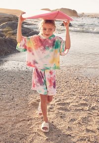 Farverigt tie-dye sweatshirt- og shortsæt i pastelfarver, med et lyserødt surfbræt holdt over hovedet, på en sandstrand.