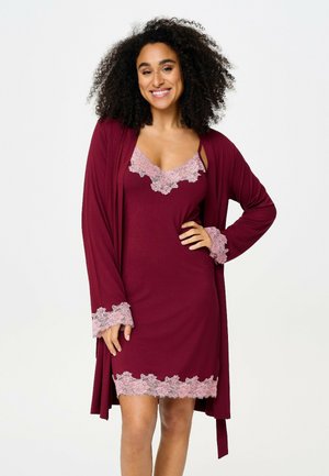 Femme souriante aux cheveux bouclés porte une chemise de nuit bordeaux et un peignoir assorti avec des bordures en dentelle rose, posant avec la main sur la hanche.