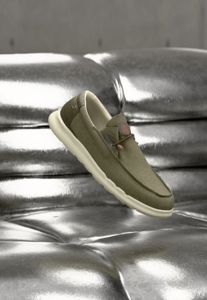 Mocassin décontracté vert olive avec semelle blanche et détails en lacets flottant au-dessus d'un canapé argenté matelassé brillant.