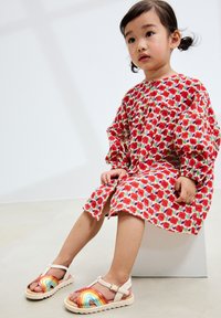 Robe florale rouge à manches bouffantes, avec des feuilles vertes sur fond blanc. Sandales à brides avec un motif arc-en-ciel et une semelle beige.