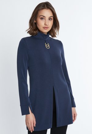 Maritiem blauwe tuniek met lange mouwen, een nepbeschuitkraag en een voorknip. Beschikt over een ronde gouden accent bij de halslijn. Gladde, rekbare stof.