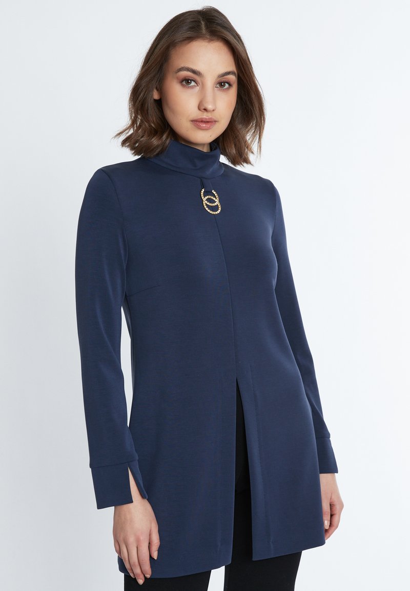 Marineblaue Langarm-Tunika mit einem Mock-Turtleneck und einem Schlitz an der Vorderseite. Verfügt über einen runden goldfarbenen Akzent am Ausschnitt. Weicher, dehnbarer Stoff.