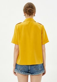 Camicia gialla a maniche corte con colletto e dettagli sulle spalle, abbinata a pantaloni shorts in denim sfilacciati. Tessuto morbido, vestibilità rilassata e linee pulite.