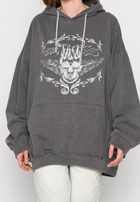 Osoba ubrana w luźną, ciemnoszaro bluzę z kapturem, na której przodzie znajduje się biały nadruk czaszki z skrzydłami, w zestawieniu z jasnymi spodniami.