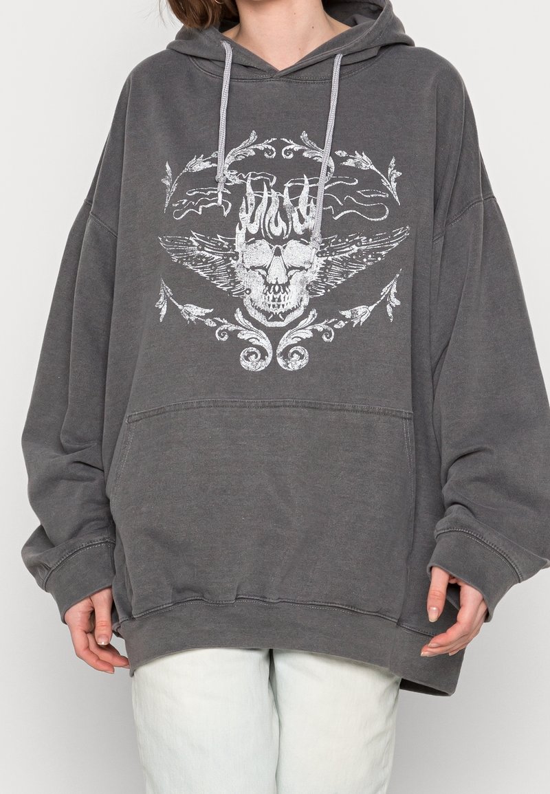 Osoba ubrana w luźną, ciemnoszaro bluzę z kapturem, na której przodzie znajduje się biały nadruk czaszki z skrzydłami, w zestawieniu z jasnymi spodniami.