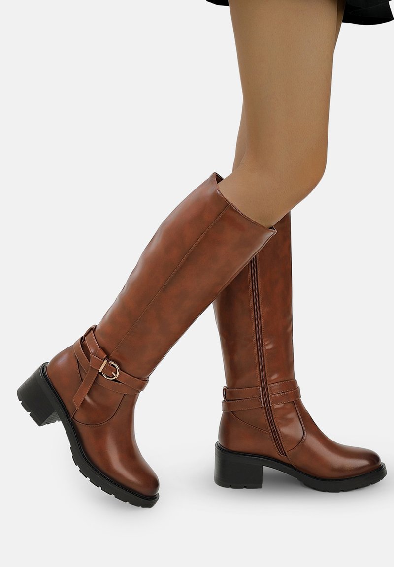 Botas marrones hasta la rodilla hechas de material sintético brillante. Cuentan con un cierre lateral, una correa decorativa con una hebilla circular y un tacón cuadrado.