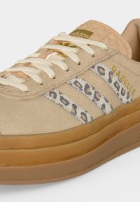 Μπεζ αθλητικό παπούτσι Adidas Gazelle με ρίγες λεοπάρ, λευκά κορδόνια, υφή στη σόλα από καουτσούκ και χρυσό λογότυπο στη γλώσσα και το πλάι.