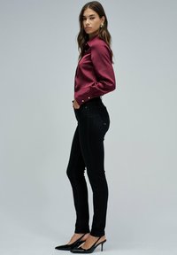 Mujer de pie de perfil, vistiendo una blusa de satén burdeos, jeans ajustados negros, zapatos de tacón negro de tipo slingback y pendientes llamativos dorados.