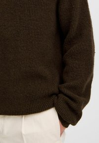Bruine gebreide trui met een geribbeld textuur en een ontspannen pasvorm. De manchette is iets omgeslagen, wat mooi aansluit bij de lichtgekleurde broek daaronder.