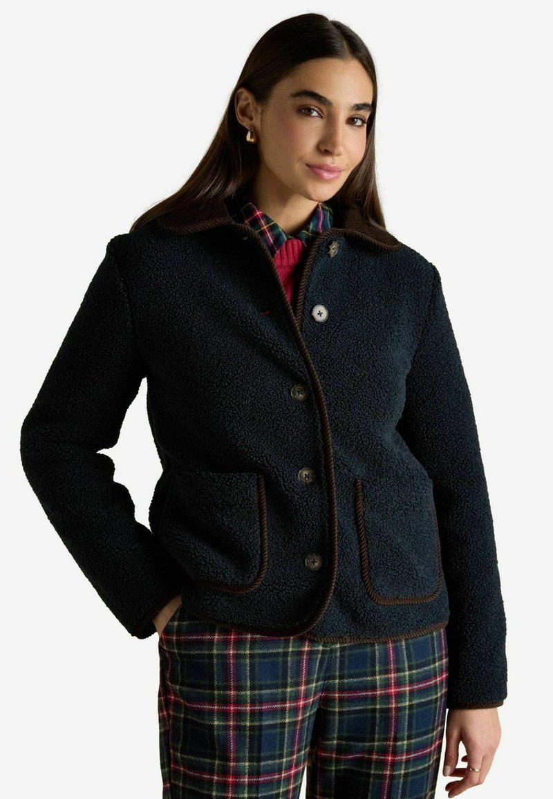 Giacca in pile blu navy con rifiniture marroni, due tasche e chiusura con bottoni. Indossata sopra una camicia a quadri e stratificata con un maglione rosso sotto.