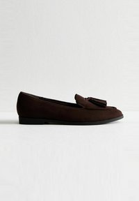 Mocasines de ante marrón con suela plana, punta redondeada y un adorno de borlas en la parte delantera. Textura suave y diseño minimalista.
