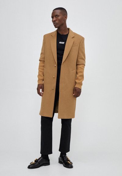 Brown wool blend coat with a tailored design, notch lapels, and three buttons. Paired with a black t-shirt and black trousers.