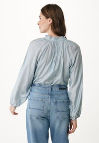 Blusa di seta azzurro chiaro con collo alto arricciato e maniche voluminose, abbinata a jeans in denim scolorito a vita alta con tasche posteriori.