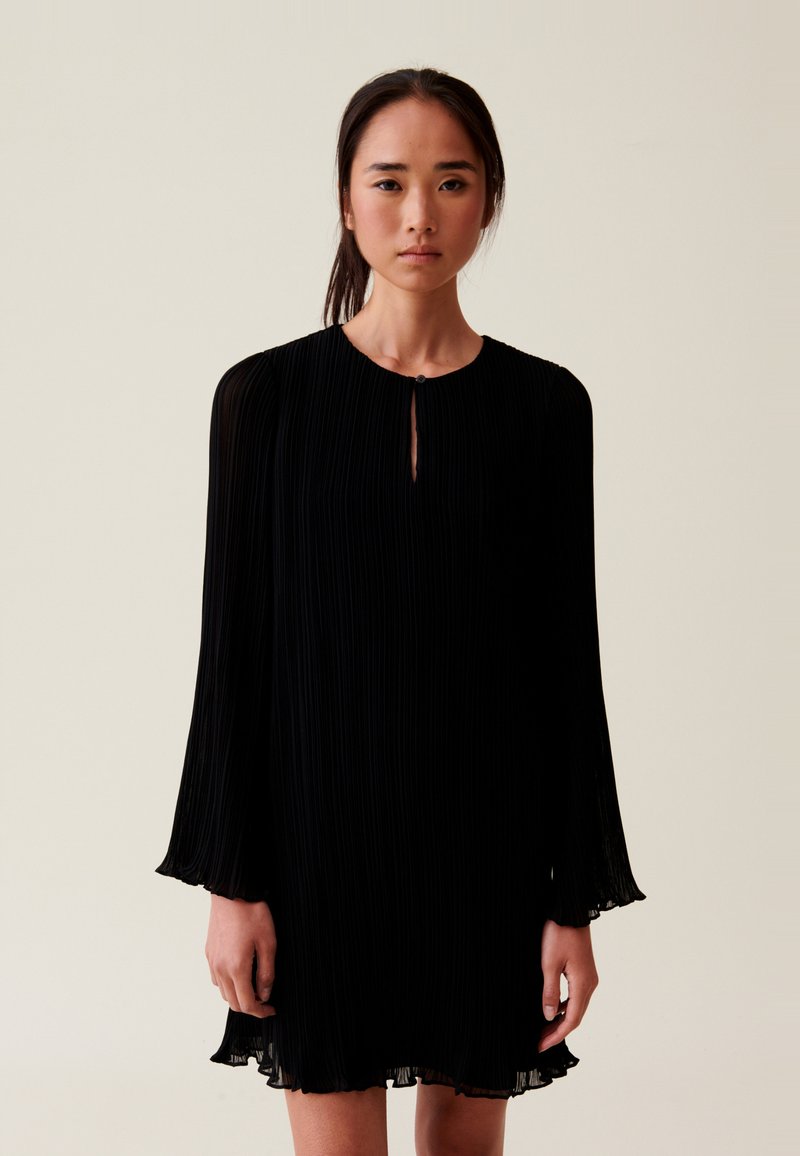 Robe noire plissée avec des manches longues, un col rond et un détail en trou de serrure à l'avant. Texture douce avec des accents transparents au niveau de l'ourlet.