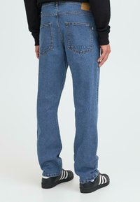 Jeans de denim de culoare albastru deschis cu croiala dreaptă, două buzunare la spate și o etichetă de marcă maro. Purtați cu pantofi negri cu dungi albe.