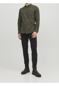 Camicia verde oliva con bottoni e taschino sul petto, abbinata a jeans neri slim-fit e stivali neri alla caviglia, su uno sfondo neutro.