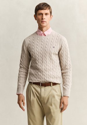 Uomo che indossa un maglione beige lavorato a trecce sopra una camicia rosa con colletto, pantaloni beige e cintura marrone, in piedi davanti a uno sfondo neutro e semplice.