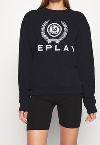 Mörkblå tröja med ribbade cuffs, prydd med en vit logotyp och lagerkransdesign; i kombination med svarta cykelshorts.