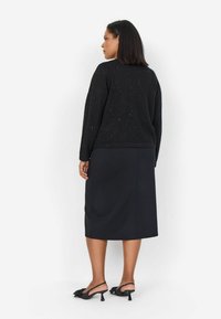 Maglione nero con sottili brillini, maniche lunghe e scollatura rotonda, abbinato a una gonna a tubino nera e décolleté nere con fiocchi.