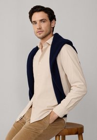 Chemise beige à boutons en dessous d'un pull en maille bleu marine drapé sur les épaules, assortie à un pantalon en velours côtelé beige, assis sur un tabouret en bois.