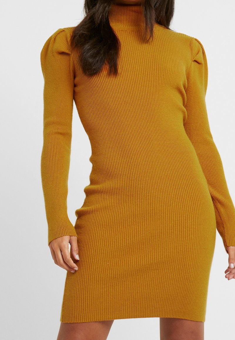Hořčicové žebrované pletené šaty s dlouhými rukávy a vysokým výstřihem. Mají jemné nabírané detaily na ramenech a přiléhavý siluetu.