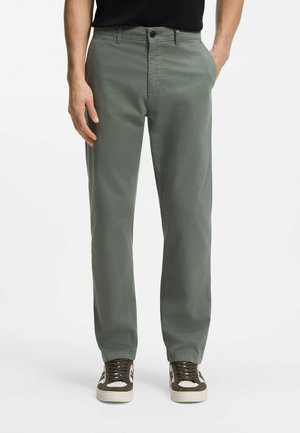 Mann trägt olivgrüne Freizeithose und braune Sneakers mit weißen Sohlen, steht mit den Händen in den Taschen vor weißem Hintergrund.