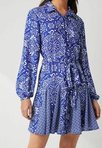 Blaues gemustertes Kleid mit Kragen, langen Ärmeln und gebundenem Taillenbereich. Besitzt florale und geometrische weiße Designs auf dem plissierten Rock.