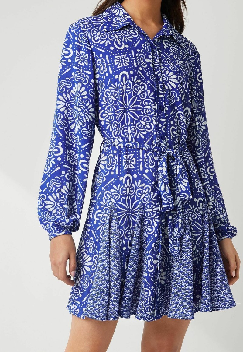 Blaues gemustertes Kleid mit Kragen, langen Ärmeln und gebundenem Taillenbereich. Besitzt florale und geometrische weiße Designs auf dem plissierten Rock.