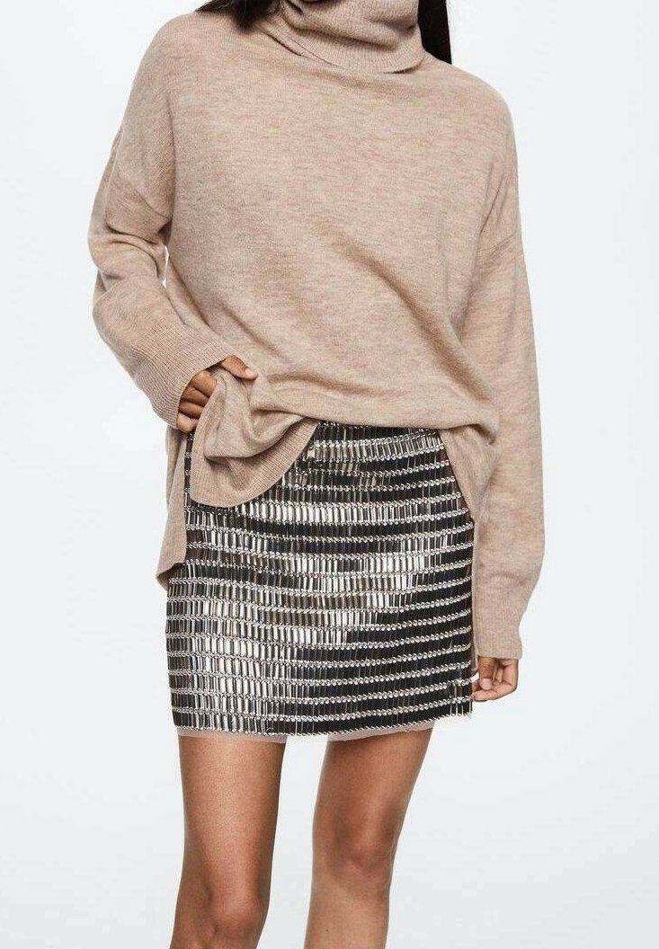Femme portant un pull à col roulé beige et une mini-jupe à motif brillant argenté et noir, debout avec une main dans la poche du pull.