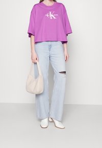 Camiseta corta morada con logo blanco, vaqueros de pierna ancha azul claro con detalles desgastados, botas blancas de tobillo y un bolso suave color crema.
