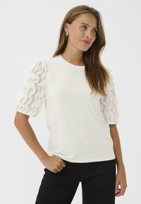 Femeie îmbrăcată cu o bluză albă cu mâneci translucide cu volane și pantaloni negri, stând cu o mână în buzunar, uitându-se ușor spre o parte.
