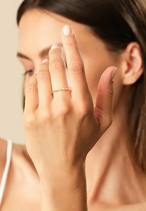 Goldring mit einem dünnen, feinen Design, das eine leichte Drehung aufweist, getragen an einer Hand mit natürlichen Nägeln vor einem neutralen Hintergrund.