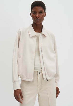 Frau trägt eine hellbeige Zip-up-Jacke über einem gerippten cremefarbenen Oberteil und cremeweißen Hosen, steht vor einem einfarbigen hellen Hintergrund.