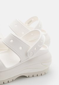 Sandale blanche avec semelle épaisse, deux brides avec des trous circulaires, semelle intérieure texturée et semelle extérieure à nervures proéminentes.