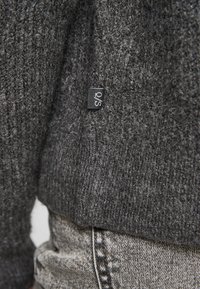 Pull en tricot gris avec une finition texturée et une petite étiquette noire, associé à un jean en denim gris clair, montrant un gros plan du tissu et des coutures.